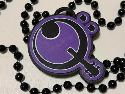A purple and black pendant necklace featuring a Q shaped guitar, displayed on a white surface with a black beaded necklace draped around it. Custom made by NickNacks for the Bass for Berry fundraiser benefiting Quintin Berry.