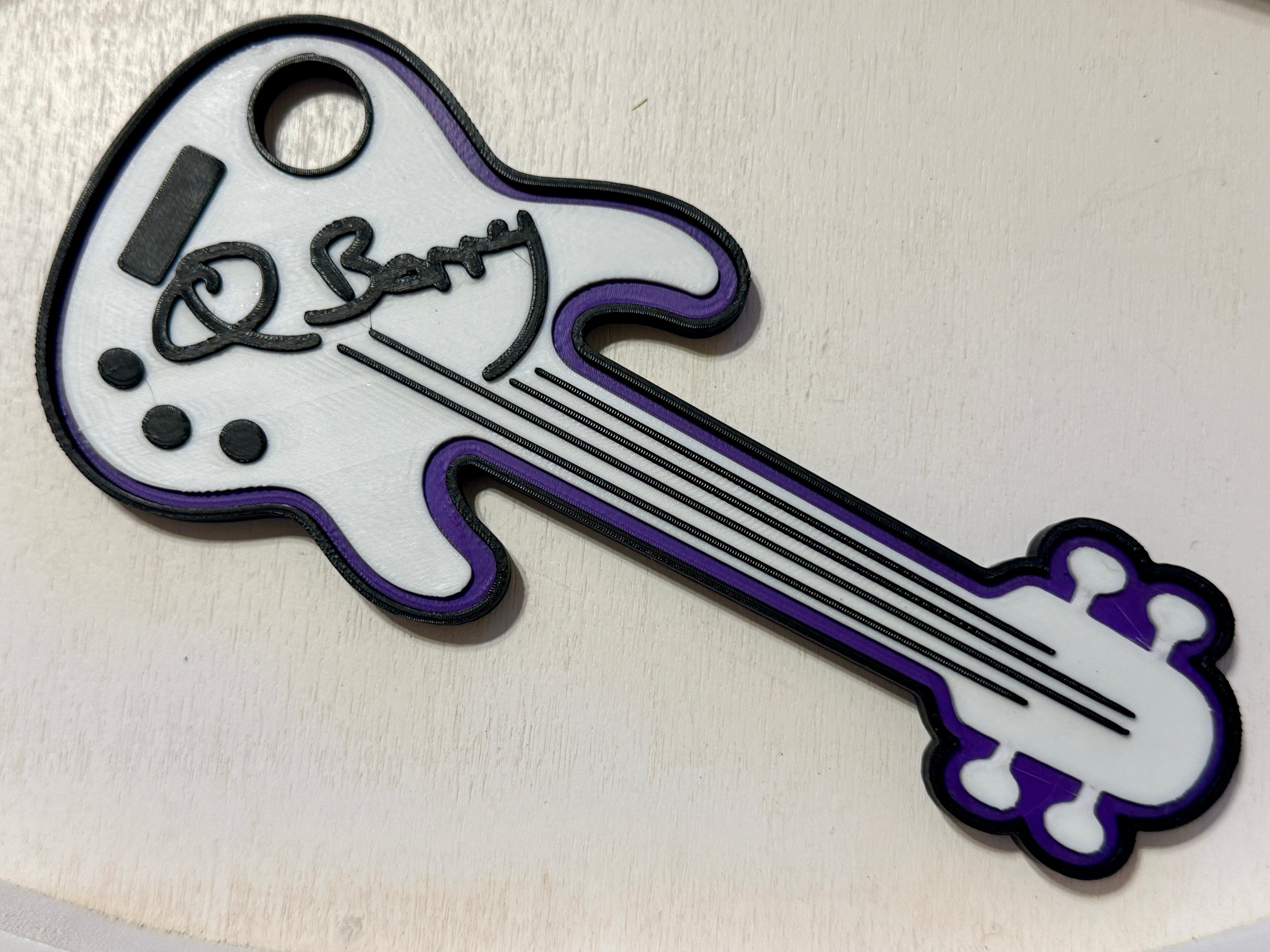 A 3D printed custom guitar-shaped figure keepsake featuring a white body with purple trim, black outlines, and a black script signature reading 'Q Barry' on the headstock area, shown on a light beige background. Custom made by NickNacks for the Bass for Berry fundraiser benefiting Quintin Berry.