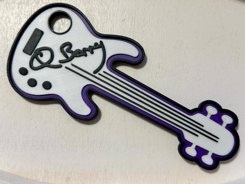 A 3D printed custom guitar-shaped figure keepsake featuring a white body with purple trim, black outlines, and a black script signature reading 'Q Barry' on the headstock area, shown on a light beige background. Custom made by NickNacks for the Bass for Berry fundraiser benefiting Quintin Berry.